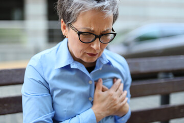A mature gray-haired woman in glasses took hold of her heart while sitting on a bench outside.