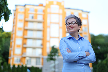 Cheerful smiling mature woman realtor with crossed arms on her chest against the backdrop of a new house.