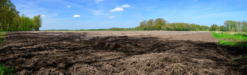 Panorama photo of a plowed farm field surrounded by forests and a blue sky.