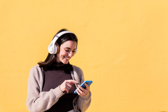 smiling young woman listening to music from phone on headphones with yellow background, concept of rhythm and positive people, copy space for text