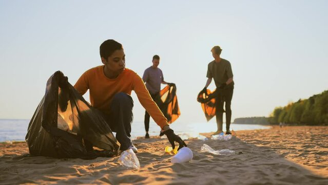 Guys volunteers cleaning sandy beach collecting trash garbage near ocean at sunset. Ecology lifestyle, environment pollution and protection. Putting on trash, rubbish, waste, junk in plastic bags.