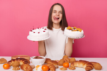 Hungry girl posing at table with sugary desserts and bakery isolated over pink background showing...