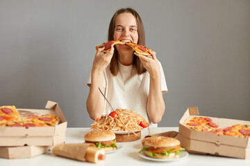 Hungry funny woman with brown hair wearing white T-shirt sitting at table among fast food isolated over gray background holding two slices of pizza eating junk food.