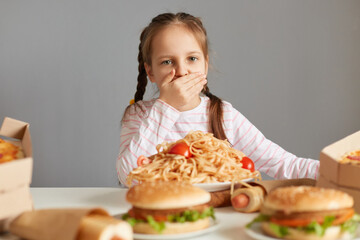 Sick ill little girl with braids sitting at table with junk food isolated over gray background feeling overating being seek feels nausea covering mouth with plam vomit.