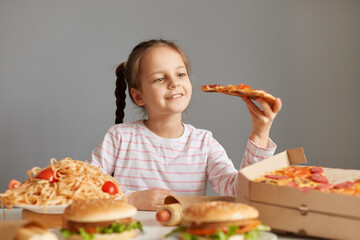 Enjoyable smiling beautiful little girl with braids sitting at table with junk food isolated over gray background holding big slice of pizza being reading to eat uhealthy dish.