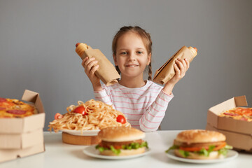 Satisfied delighted little girl with braids sitting at table with fast food isolated over gray...