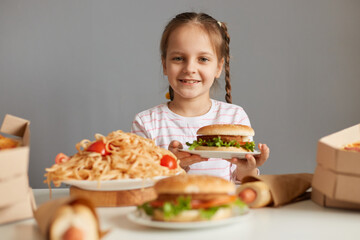 Cute positive smiling little girl with braids sitting at table with fast food isolated over gray background looking at camera with happy expression eating tasty hamburger.