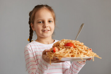 Cute smiling little kid girl with pigtails eating spaghetti holding big plate with pasta looking at...