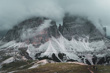 Hiking path with two people walking along a trail in front of a massive mountain with a hut in front in the Dolomites, Italian alps with a storm approaching