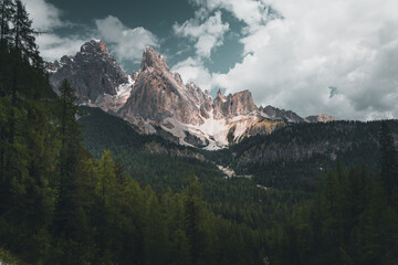 Mountain sticking out of a forest in the middle of the italian alps with the sun shining on it and clouds in the background
