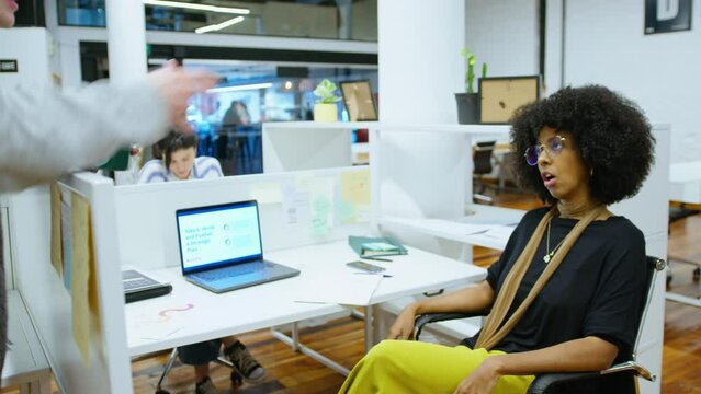 Young man leaning on cubicle wall, holding coffee mug, discussing business with African-American female colleague during workday in open space office