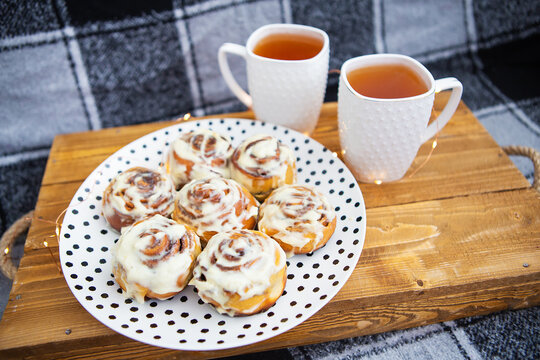 Two cups of black tea stand on a wooden tray on the sofa with a black and white checkered plaid. Fresh and fragrant cinnamon rolls close up lie on a plate with polka dots, beautiful morning