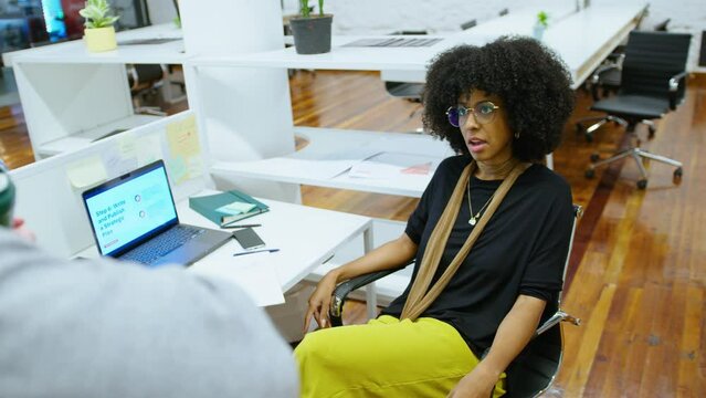 Young African-American businesswoman sitting at workplace and talking with male colleague over cubicle wall in modern open space office