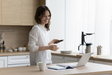 Pretty young woman using modern wireless devices spend time in domestic kitchen. Female freelancer...