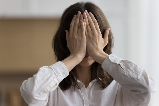 Close up head shot portrait young woman hides her face behind palms feels ashamed, shy or afraid pose indoor. No see, ignoring, dont listen to slander, gossips, try to cope with emotional breakdown