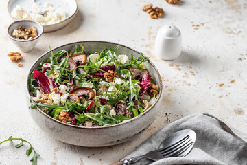 Green salad with feta cheese, arugula, mushrooms, walnut in ceramic bowl on textured background....