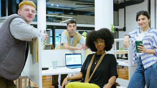 Young multi-ethnic coworkers in casual wear posing together on camera, smiling and having coffee break during workday in modern open space office with cubicles