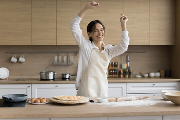 Happy young attractive woman dancing in kitchen while cooking homemade pizza, female in apron moving to favourite music, feels carefree, enjoy pastries preparation on weekend at home. Culinary, hobby - obrazy, fototapety, plakaty