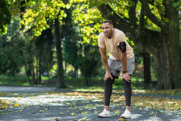 Tired man resting in park after jogging and fitness class, hispanic man using phone listening to...