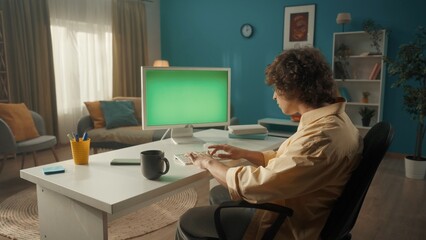 Fototapeta na wymiar A young curly guy is working on a computer with a green screen. A man sits at a table with a computer, and there is a cup of coffee next to him. Back view. Advertising area, workspace mock up.