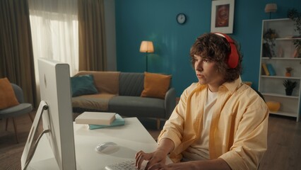 A young serious, focused guy in red wireless headphones sits at a computer. A young guy plays an online game, works, watches videos, listens to music. Close up.