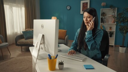 A young Asian woman with a serious look sits at a computer with a smartphone to her ear. A woman is talking on the phone while working at a computer. The concept of technology, communication.