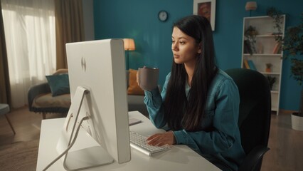 An Asian woman sits at a computer. In one hand, a woman holds a cup of coffee, the other is typing...