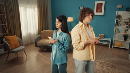 A young distressed married couple is typing messages on their smartphones. Asian woman and man stand with their backs to each other, thinking about the message text.