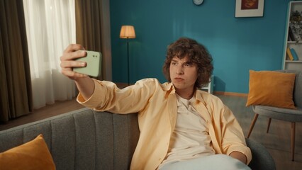 A young man takes a selfie on a smartphone. Portrait of a curlyhaired man on the sofa in the living room with a phone in his hands close up. © kinomaster