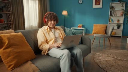 A young man is sitting on a couch wearing red wireless headphones with a laptop on his lap. A man is typing a text, a message, chatting, listening to music, playing online games.