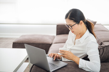 adult woman doing research work for her business. Asian woman sitting on sofa relaxing while browsing online shopping website. Happy lady browsing through the internet during free time at home.