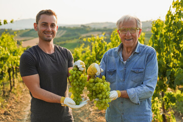 Grandfather and grandson smiling on camera in their vineyard while holding fresh grapes -...