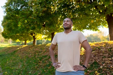 Fotobehang Smiling sportsman resting in the park on a run, man breathing fresh air enjoying an active lifestyle, hispanic man exercising. © Liubomir