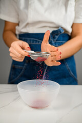 Confectioner sprinkling red powder over empty plastic cooking bowl on kitchen table