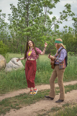 Couple of young people in hippie style. A girl is dancing, a guy is playing a guitar on a country road on a sunny day
