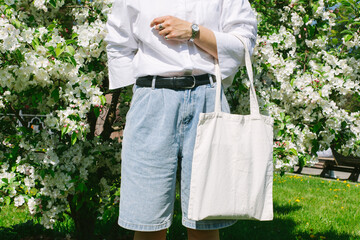 Woman hand holding eco bag. Female hold white canvas textille mesh zero waste shopper on city street, green apple tree flowers background. Blank mock up tote bag, place for text design logo. 