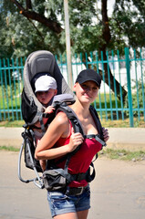 Obraz premium A young mother with a small child in a carrier bag on her back. Hiking with a small child in summer. Vertical photo. Side view