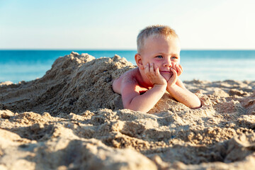 A little boy lies buried in the sand on the seashore on a bright sunny day. Close-up. Tourism, travel and recreation.