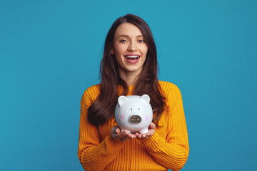 Fototapeta na wymiar Cheerful young girl in casual orange sweater smiling and holding white piggy bank with lots of money isolated over blue background