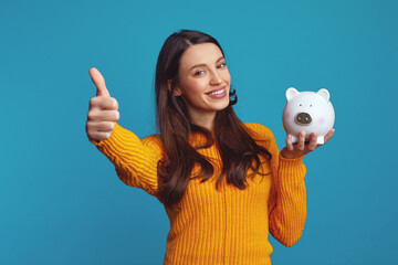 Happy young woman in casual orange sweater holding white piggy bank with lots of money, showing...