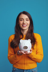 Vertical photo of happy young woman in casual orange sweater holding white piggy bank with lots of...
