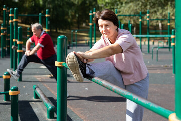 Fototapeta premium Positive elderly woman doing leg stretching on outdoor sports ground