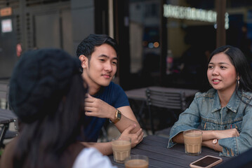 A guy in a blue shirt gazing far while sitting with lady pals, one in a denim jacket and the other is wearing a white long sleeves and a beret. Window glass, table and chairs in the background.