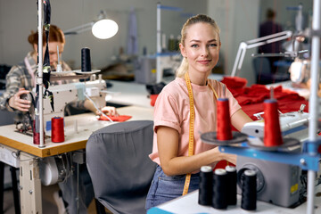 Dressmakers women sewing clothes on sewing machine in factory together, at work. Attractive ladies enjoy tailoring, making clothes, blonde caucasian craftswoman looking at camera smiling