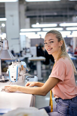 young nice caucaian blonde textile factory worker on production line using modern sewing machine, at workshop. hardworking woman engaged in sewing or tailoring,looking at camera smiling - obrazy, fototapety, plakaty