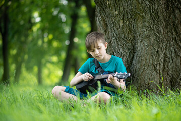 a boy plays a small guitar, a black ukulele on a bench in the park