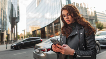 Fashion beautiful woman in stylish clothes with a black leather jacket walks and uses a smart phone in a modern city