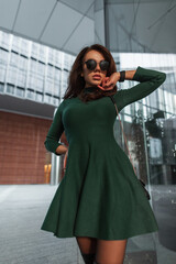 Fashion beautiful chic lady with stylish sunglasses in a fashionable green dress with handbag walks in the city near the modern business glass office building