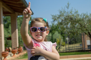 Obraz premium Image of adorable smiling little girl with sunglasses in home garden holding index finger and arm raised. Little girl playing model. 