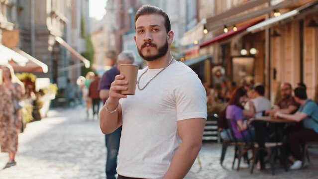 Happy bearded young man enjoying morning coffee hot drink and smiling outdoors. Relaxing, taking a break. Guy walking in urban city sunshine street, drinking coffee to go. Town lifestyles outside